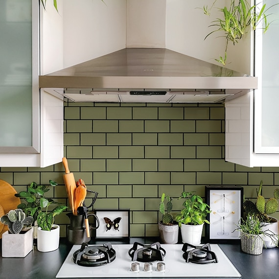 Closeup of white gas stove top on black countertop holding houseplants, green subway tile backsplash with black grout, white cabinets, and metallic vent hood.