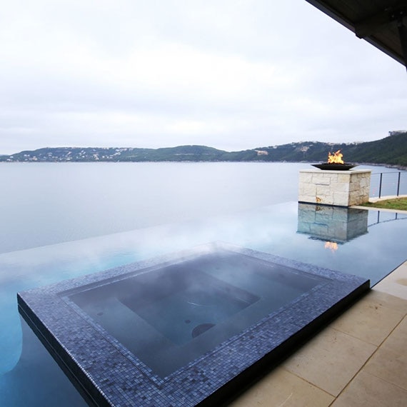 Infinity pool with hot tub covered with dark blue mosaic tile, fire pit stone column, and view of large lake and hillside in the background.
