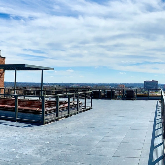 Rooftop with 2CM, gray stone look pavers, metal rails, air conditioner units, and a blue sky background with white fluffy clouds.