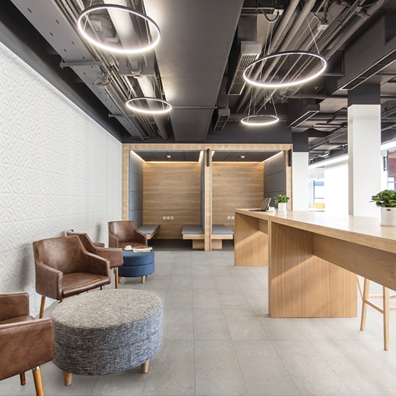 Office breakroom with floor tile that looks like gray stone, feature wall with white textured tile, brown leather chairs, long wood counter, round lighting hanging from black metal ceiling.