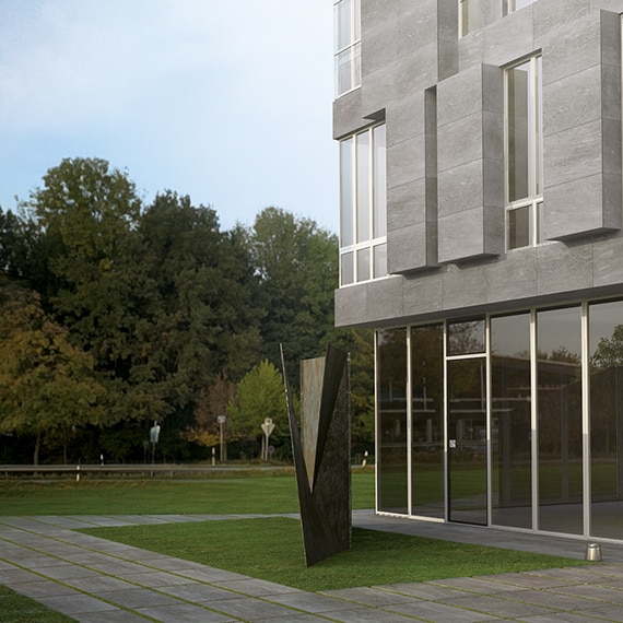Exterior of office building with glass and gray stone look cladding, walkway with matching 2cm porcelain pavers set in grass, and gray metal sculpture.