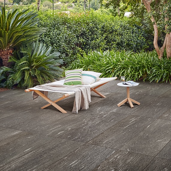Backyard patio with wooden & linen cot & side table on dark gray tile pavers that look like stone and surrounded by foliage.