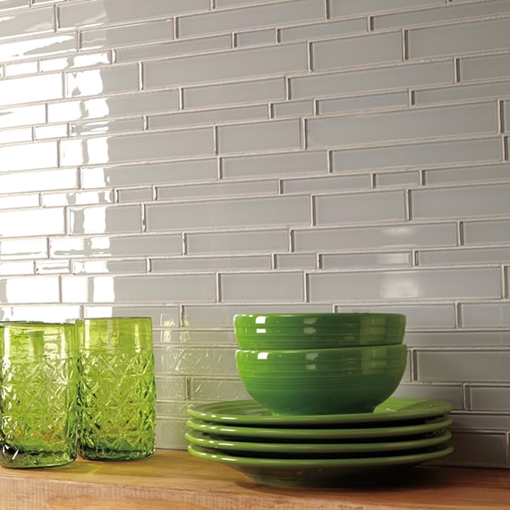 Closeup of gray glass mosaic tile backsplash with floating wood-grain shelf holding bright green plates, bowls, and drinking glasses.