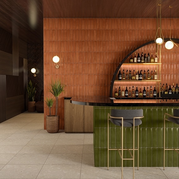 Restaurant bar with orange glossy wall tile, bottles of alcohol on brass shelves, black porcelain slab countertops, bar front with green glossy tile and beige terrazzo look floor tile.