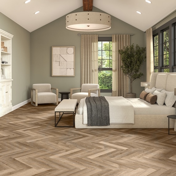 Transitional bedroom with LVT wood look floor tile in herringbone pattern, muted green walls, white platform bed, upholstered bench and side chairs.
