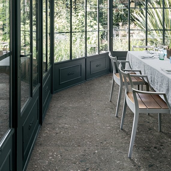 Restaurant dining room with skylight, gray floor tile that looks like terrazzo, table and chairs surrounded by windows with a view of a garden.