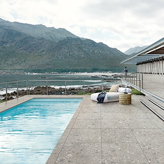 Pool with stone look porcelain tile deck, lounge chair and table with mountain and rocky beach in the background.