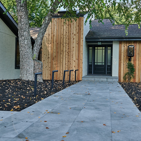 Home entrance with black & glass door, white stucco, wood panel accents, walkway with light gray 2CM porcelain pavers, and large tree.