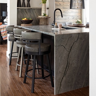 Kitchen with gray natural quartzite island with sink and waterfall countertop, gray leather and metal bar stools, pendant lighting.