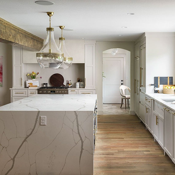 Renovated kitchen with white with gray veining quartz countertops and waterfall island, white cabinets, brass & glass pendants, and ceiling wood beam.