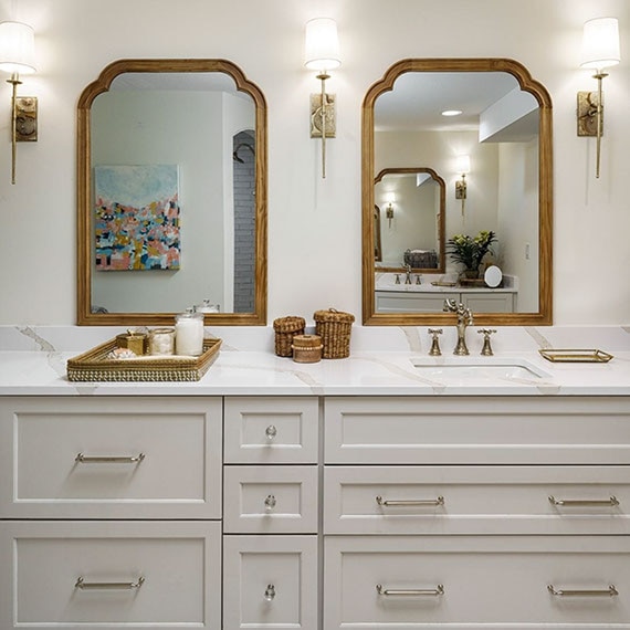 Renovated bathroom with marble look white quartz vanity countertops, antique brass faucet, framed mirrors, and white cabinets with antique brass accents.