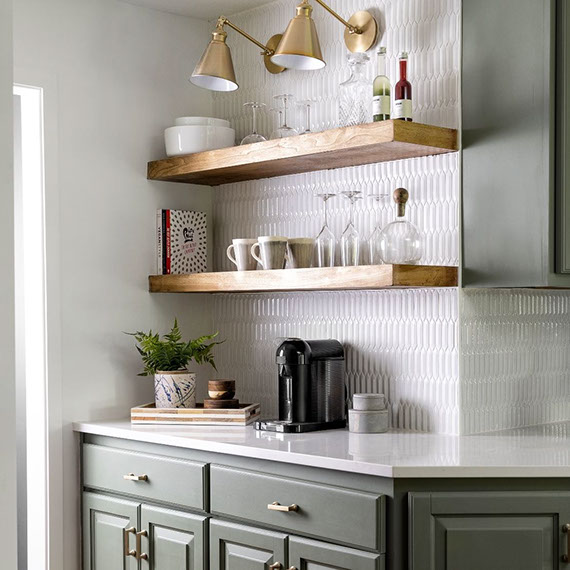 Renovated kitchen with white quartz countertops, white picket mosaic tile backsplash, olive cabinets, floating woodgrain shelves, and brass sconces.