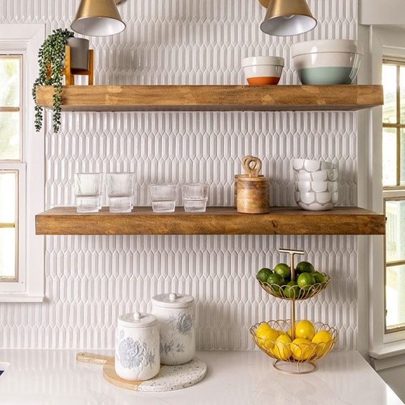 Renovated kitchen with white quartz countertops, white picket mosaic tile backsplash, floating woodgrain shelves, and brass sconces.