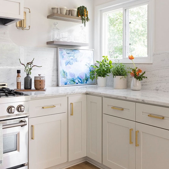 Renovated kitchen with gray marble backsplash and countertops, floating shelves, and off-white cabinets with brass accents.