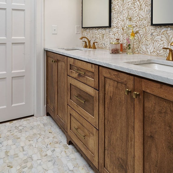 Renovated bathroom with beige & gray marble mosaic floor tile, gray marble counter on a wood grain vanity, framed mirrors on white & beige marble mosaic backsplash.