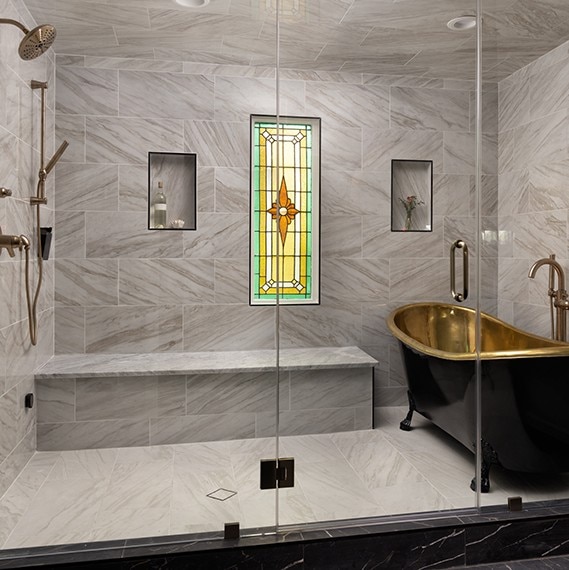 Renovated bathroom wet room with stain-glass window, black & gold soaker tub, white & gray marble look shower wall, floor tile and niches.