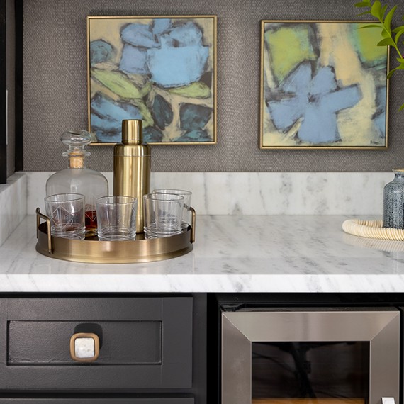 Renovated dry bar with decanter, shaker & highball glasses sitting on white & gray marble countertop, black cabinets, wine refrigerator, and blue & yellow paintings.