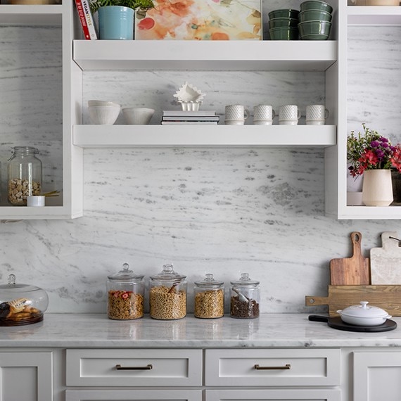 Renovated kitchen with white & gray marble backsplash and countertops, white floating shelves and lower cabinets, and glass canisters containing grains.