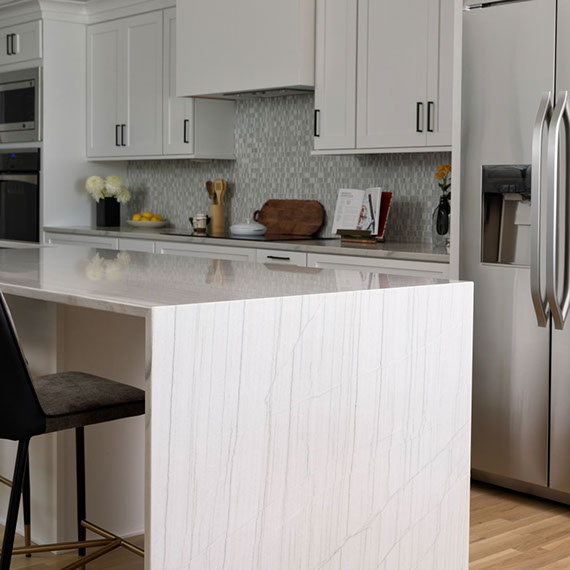 Renovated kitchen with gray natural quartzite countertops and waterfall island, beige & white glass mosaic backsplash, white cabinets and floating shelves.