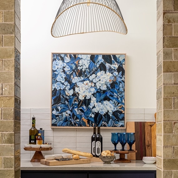 Renovated pantry with glossy white tile backsplash, blue flower painting, and cage pendant over a taupe quartz countertop.