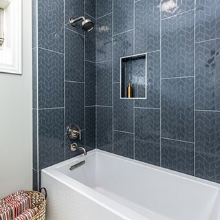Closeup of bathtub/shower combination of blue chevron shower wall tile, brushed silver showerhead & faucet, and shower shelf holding a brass liquid dispenser.