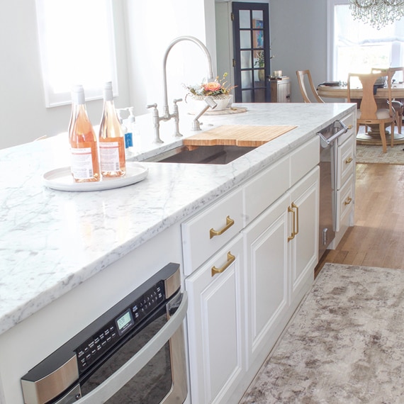 Renovated kitchen with white & gray marble waterfall island with sink, white cabinets, and brushed brass faucet.