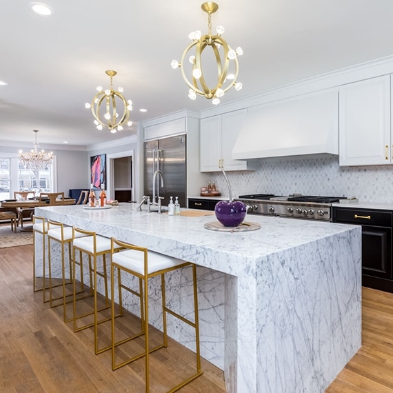 Kitchen with marble mosaic backsplash, white quartz countertops, marble waterfall island with sink, white cabinets, and brushed brass pendant lights.