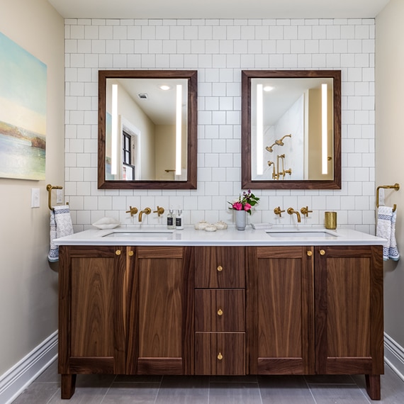Remodeled bathroom with white square ceramic tile backsplash, natural wood vanity with dual sinks, wood framed mirrors, and brushed brass wall-mounted faucets.