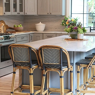 Renovated kitchen with heavily veined gray quartzite backsplash, island, and countertop, wicker bar stools, gray cabinets, and sink in front of large picture window.