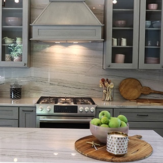 Renovated kitchen with heavily veined gray quartzite backsplash, island, and countertop, gray cabinets, and stainless steel gas stove.