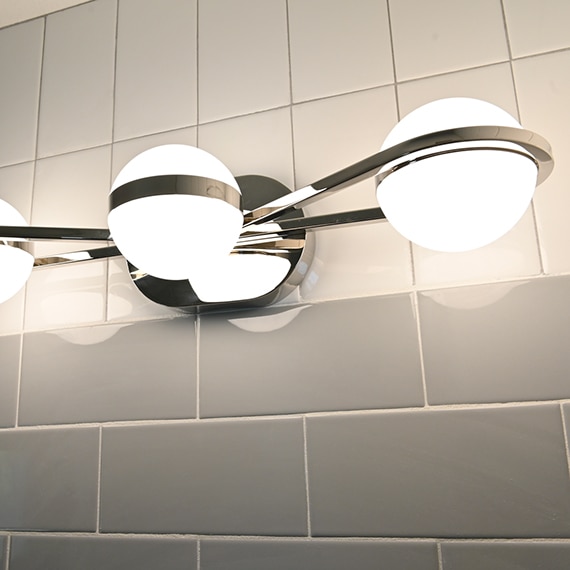 Closeup of bathroom wall with gray subway tile backsplash, white vertical tile trim, and three globe light polished silver wall sconce.