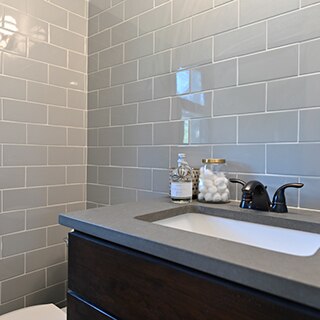 Closeup of bathroom vanity with gray subway tile backsplash, gray quartz countertop, black faucet, under-mount sink, and glass container of cottonballs.