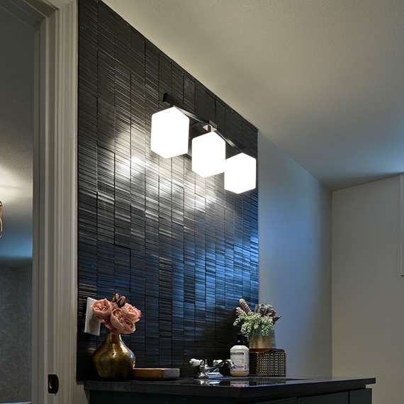 Bathroom with backsplash of black metallic tile, black quartzite vanity countertop, triple dangling sconces, and small brass vase holding pink roses.