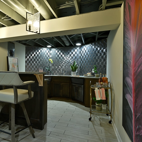 Basement bar with silver textured geometric wall tile that looks like metal tile, floor tile that looks like wood flooring, and beige leather bar stools.