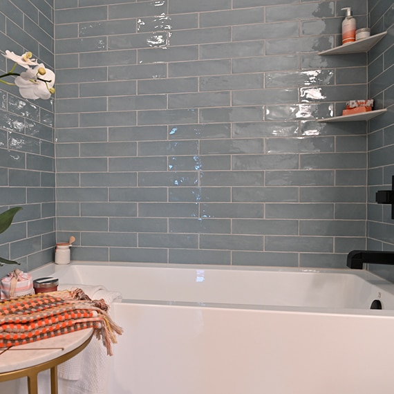 White orchids next to shower of blue ceramic subway tile with built-in corner shelves, white bathtub and matte black faucet mounted on the shower wall.