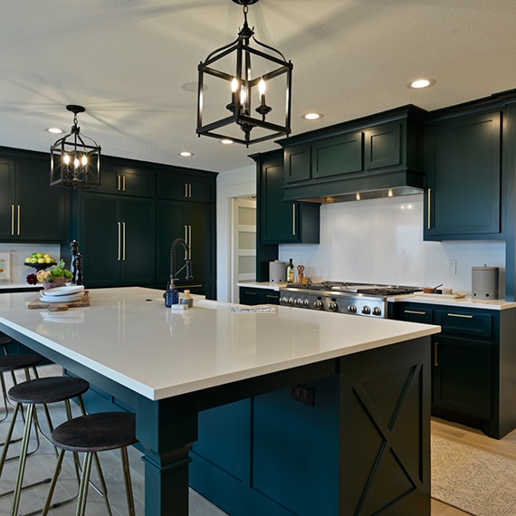 Remodeled kitchen with white quartz countertops and backsplashes, dark green cabinets with brass hardware, and black cage pendants over island.