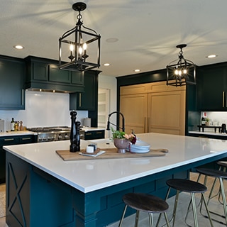 Remodeled kitchen with white quartz countertops and backsplashes, dark green & natural wood cabinets with brass hardware, and black cage pendants over island.