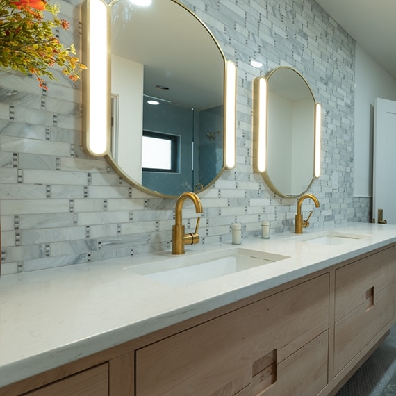 Bathroom quartz countertop with double sinks, brushed brass faucets, marble mosaic backsplash, and lighted vanity mirrors.