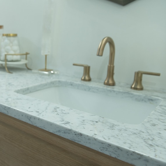 Undermount bathroom sink with brushed brass faucet, white with gray veining marble countertop, natural wood cabinet.