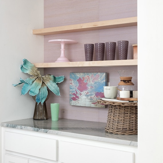 Coffee nook with natural quartzite countertop, pink wall paper, natural wood floating shelves holding purple glassware and pink cake stand.