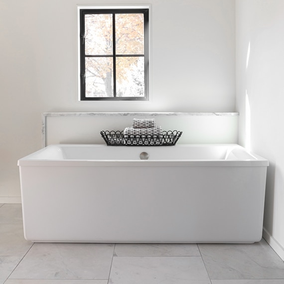 Freestanding bathtub with white quartz & marble trim backsplash, black basket holding gray rolled towels, 24x24 marble tile flooring.
