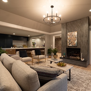 Open concept kitchen/family room with large quartz island, dark gray mosaic fireplace, coffee table, tan overstuffed sofa & side chairs.
