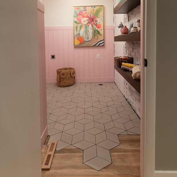 Laundry room with pink shiplap wainscoting, painted brick wall with floating wood shelves, 3D cube porcelain tile flooring, and painting of colorful flowers.