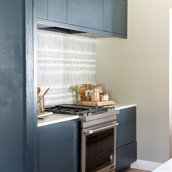 Stainless steel gas stovetop & oven, blue cabinets, gray wallpaper, and glass jars on white marble look quart countertop.