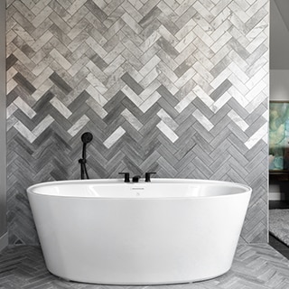 Freestanding soaking tub in front of chevron backsplash of white & gray marble tile blended with gray limestone tile, and matte black faucet.