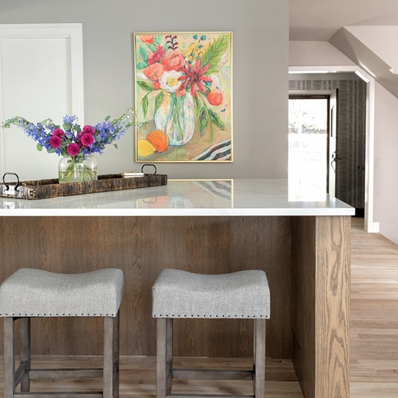Kitchen island with a bouquet of pink & purple flowers on a white quartz countertop, natural wood base, with gray bar stools.