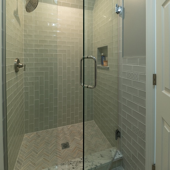 Shower with light green tile, marble niche & threshold, herringbone mosaic gray/tan marble shower floor tile, and glass door.