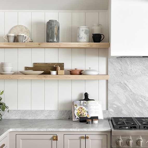 Kitchen with white marble countertops and backsplash, wood shelves with dishes, silver stove, white walls, and off white cabinets.