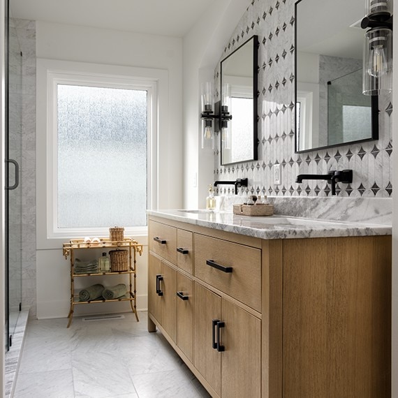 Bathroom sideview with light brown wood cabinets on his and hers sinks, white marble floor, white marble look mosaic on wall, and white marble slab on the countertops.
