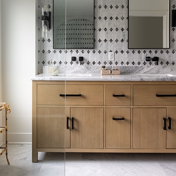 Bathroom with light brown wood cabinets on his and hers sinks, white marble floor, white marble look mosaic on wall, and white marble slab on the countertops.